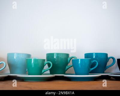 Set di grandi e piccole tonnellate verdi blu di tazze da caffè, tazze in ceramica con piattini stand in fila su ripiano di legno isolato su sfondo bianco con copia sp Foto Stock