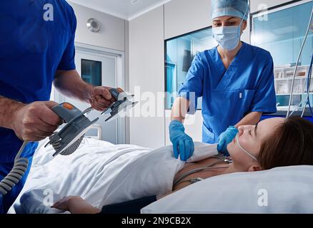 team medico che esegue una rianimazione con il defibrillatore su una paziente incosciente sdraiata nel reparto ospedaliero. RCP in terapia intensiva Foto Stock