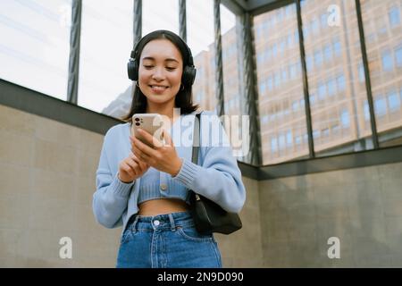 Giovane donna asiatica sorridente e utilizzando il cellulare mentre si è in piedi in casa Foto Stock
