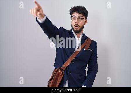 Uomo ispanico con barba che indossa abiti da lavoro puntando con il dito sorpreso davanti, bocca aperta stupita espressione, qualcosa sul fronte Foto Stock