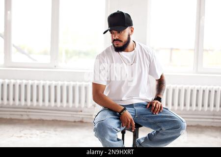Autentico uomo portatore seduto su sfondo bianco loft, guardando lontano, indossando casual T-shirt bianca, berretto da baseball e jeans, stile di vita Foto Stock