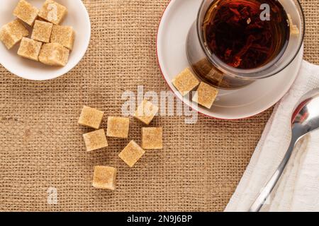 Diversi cubetti di zucchero di canna con un bicchiere di tè nero e un cucchiaio di metallo su un panno di iuta, macro, vista dall'alto. Foto Stock