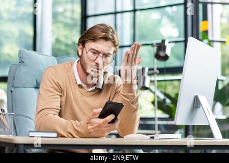 Arrabbiato giovane programmatore maschio, freelancer seduto in ufficio al tavolo, tenendo il telefono in mano, guardando in modo orribile. Si è verificato un problema, cattive notizie, messaggi, controllare l'account. Foto Stock