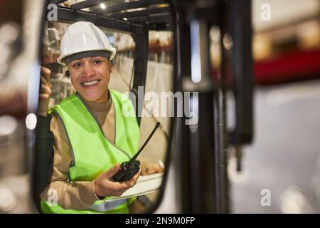 Ritratto di una giovane donna sorridente che indossa un elmetto durante il funzionamento del carrello elevatore a forche in magazzino, spazio copia Foto Stock