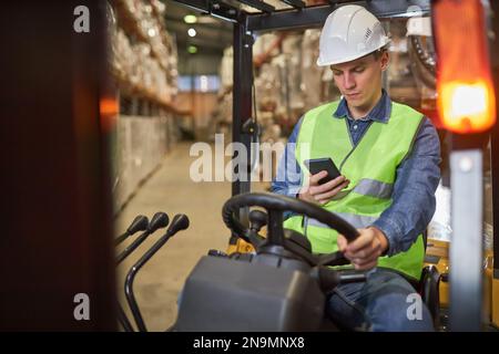 Ritratto di un giovane lavoratore che guida un carrello elevatore a forche in magazzino e utilizza uno smartphone Foto Stock