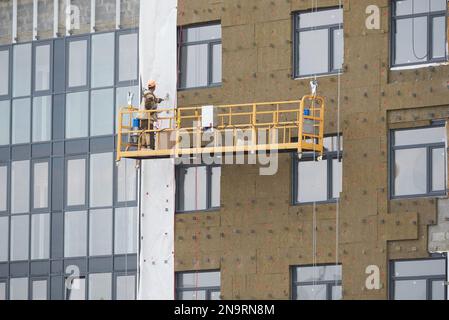 Il costruttore lavora in una speciale piattaforma sospesa e isola il muro esterno di un condominio Foto Stock