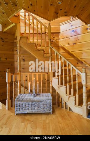 Vecchio tavolo in vimini taupe con candele accese di fronte alla scala in legno di pino che conduce al seminterrato e al piano superiore dal soggiorno in una capanna di legno. Foto Stock