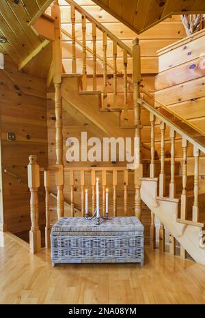 Vecchio tavolo in vimini taupe con candele accese di fronte alla scala in legno di pino che conduce al seminterrato e al piano superiore dal soggiorno in una capanna di legno. Foto Stock