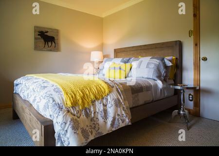 Letto queen size su cornice di legno marrone chiaro e comodino cromato nella camera da letto degli ospiti nel seminterrato all'interno di una casa in legno di pino bianco orientale. Foto Stock