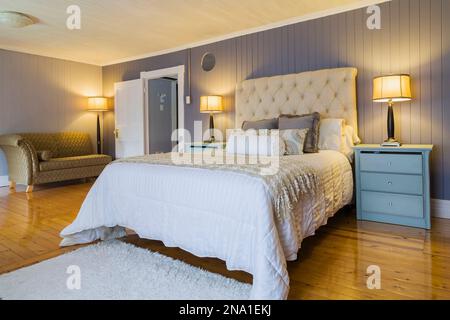 Letto queen size con copriletto bianco, testiera in tessuto beige, tavoli con estremità blu chiaro e recamier rivestito in oro e grigio nella camera da letto principale della vecchia casa. Foto Stock