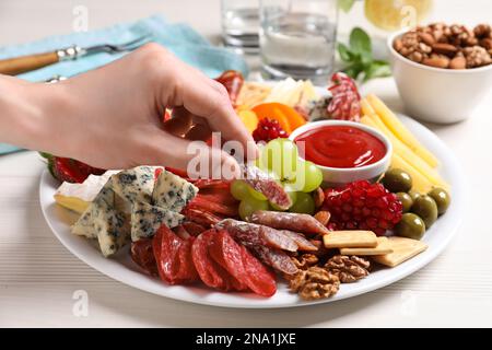 Donna che prende salsiccia dal piatto con antipasti diversi su tavolo di legno bianco, primo piano Foto Stock