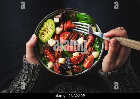 Insalata sana fatta in casa con pomodori, cubetti di formaggio, noci pecan, avocado, spinaci e rucola. Pasto vegetariano verde in una ciotola nelle mani della ragazza su un dar Foto Stock