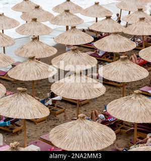 Turisti in spiaggia in sdraio poste in file sotto gli ombrelloni di paglia; Budva, comune di Budva, Montenegro Foto Stock