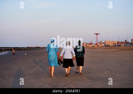 Donne ebraiche ortodosse che camminano sulla spiaggia di Coney Island dopo l'alba; Coney Island, New York, Stati Uniti d'America Foto Stock