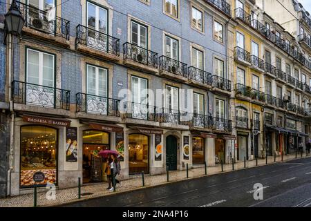 Negozi e appartamenti lungo una strada con pedoni in possesso di un ombrello in una giornata di pioggia; Lisbona, Regione di Lisboa, Portogallo Foto Stock