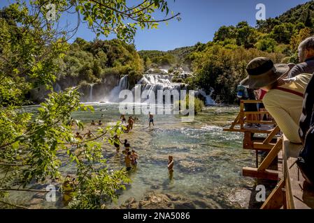 Persone che nuotano nel fiume Krka presso le cascate di Krka con turisti in piedi sul ponte che si affacciano sul Parco Nazionale di Krka; Croazia Foto Stock