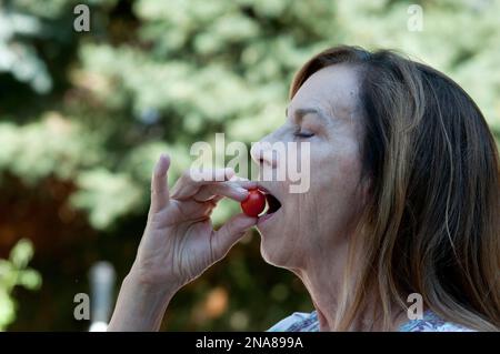 Donna anziana che mangia un pomodoro fresco di ciliegia (MR) Foto Stock