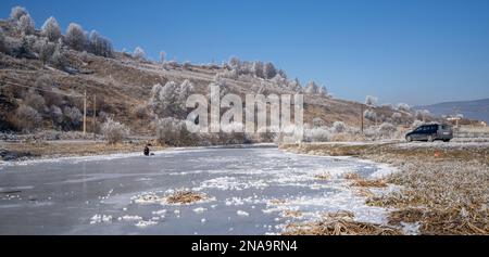 Uomo che pesca attraverso il buco nel ghiaccio del fiume ghiacciato nella Transilvania rurale in inverno, Romania; Transilvania, Romania Foto Stock