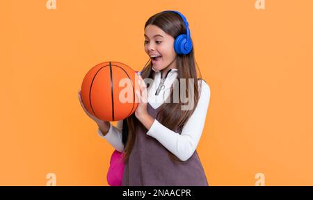 il bambino ha sorpreso le cuffie con lo zaino della scuola e la palla da basket su sfondo giallo Foto Stock