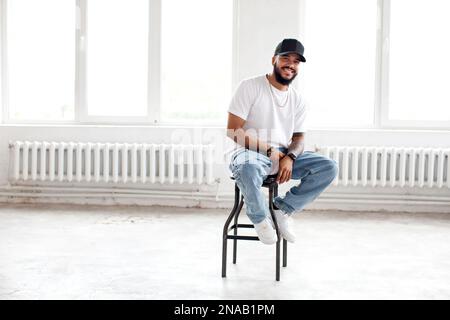 Autentico Ritratto felice uomo portatore seduto in bianco sfondo loft, sorridente, indossando casual T shirt bianca, berretto da baseball e jeans, lifestyle Foto Stock