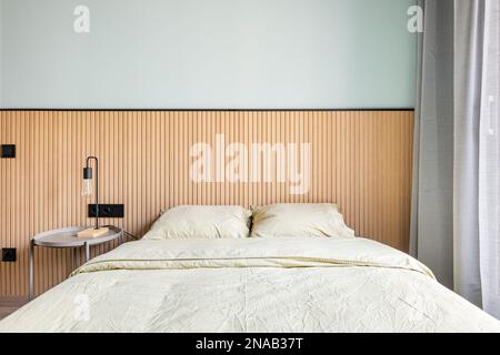 Dalla finestra, la luce del giorno illumina la camera da letto con una parete in menta e un decor in legno. Primo piano di un letto matrimoniale con biancheria beige. Tavola rotonda di Foto Stock