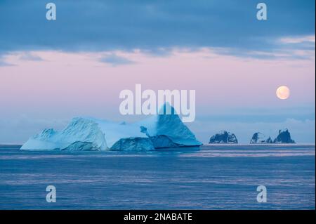 La luna piena sorge su iceberg e isole montuose nello stretto di Gerlache al largo della costa della penisola antartica; Antartide Foto Stock