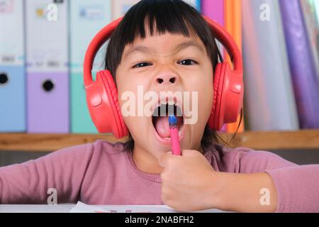 Allegra bambina in cuffie che cantano e disegnano con pastelli su carta seduti al tavolo nella sua stanza a casa. Creatività e sviluppo di fine Foto Stock