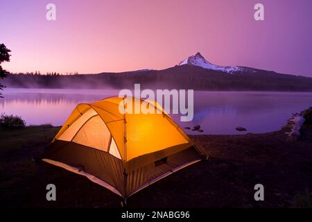 Tenda illuminata sul bordo dell'acqua di un grande lago all'alba con il Monte Washington sullo sfondo; Oregon, Stati Uniti d'America Foto Stock