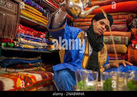 Un uomo di un villaggio berbero, vestito con abiti tradizionali, versa tè marocchino in un negozio di tappeti a Erfoud, Marocco; Erfoud, Marocco Foto Stock