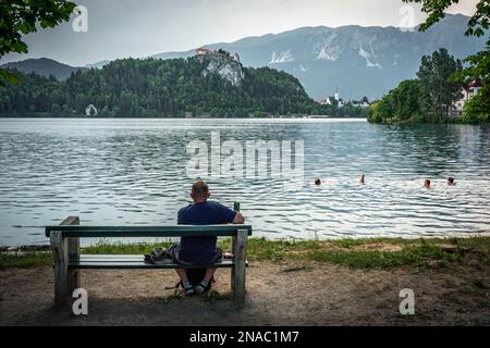 Vista posteriore di un uomo rilassato seduto su una panchina accanto ad un lago di Bled con una lattina di birra in mano, guardando la gente nuotante e il castello di Bled Foto Stock