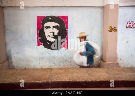 Un uomo porta un carico vicino a un dipinto di che Guevara sul muro di un mercato agricolo cubano; Cienfuegos, Cuba Foto Stock