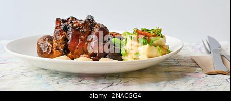 Ginocchio succoso di maiale al forno con purè di patate e verdure sul piatto, posate sul tavolo, primo piano. Foto Stock