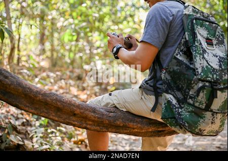 Immagine ritagliata di un escursionista asiatico maschio o viaggiatore con zaino e fotocamera retrò siede su una vite di legno, riposandosi mentre trekking nella foresta. Foto Stock