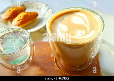 Chiudiamo una tazza di caffè latte con torte della Madeleine sfocate sullo sfondo Foto Stock