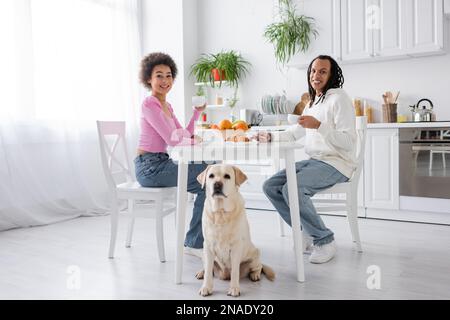 Sorridente coppia afroamericana che tiene il caffè vicino labrador in cucina Foto Stock