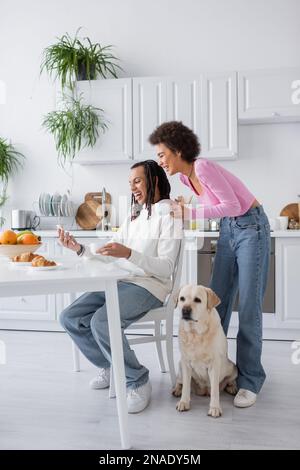 Allegra coppia afroamericana utilizzando smartphone e tenendo il caffè vicino labrador in cucina Foto Stock