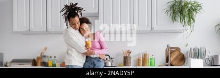 Coppia afro-americana sorridente con succo d'arancia che abbraccia in cucina, banner Foto Stock