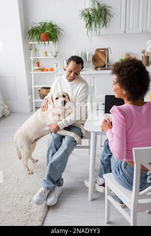 Donna afro-americana sorridente che tiene il caffè vicino al ragazzo con labrador in cucina Foto Stock