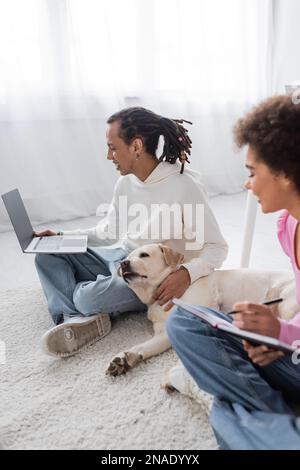 Vista laterale di labrador, sdraiato vicino ai freelance afro-americani che lavorano sul tappeto a casa Foto Stock