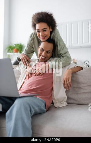 Donna afroamericana che abbraccia il ragazzo tatuato vicino al laptop sul divano a casa Foto Stock