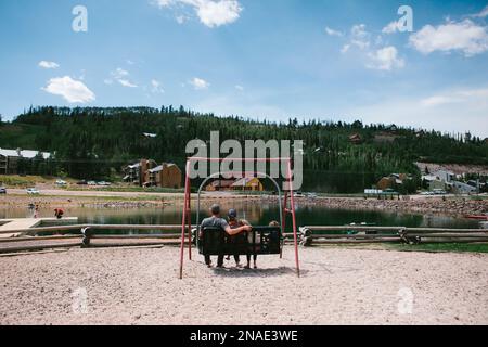 La famiglia si siede sulla panca che oscilla insieme al lago di montagna in estate Foto Stock