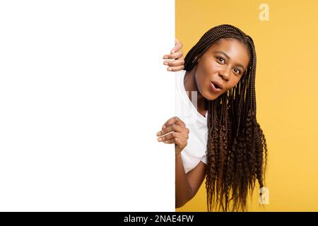 Divertente stupita donna africana si scaccia da dietro vuoto banner bianco, nascondendosi dietro enorme poster di carta, ragazza con pigtail sorpreso con l'affare, la pubblicità, mock up, copia spazio, isolato sul giallo Foto Stock