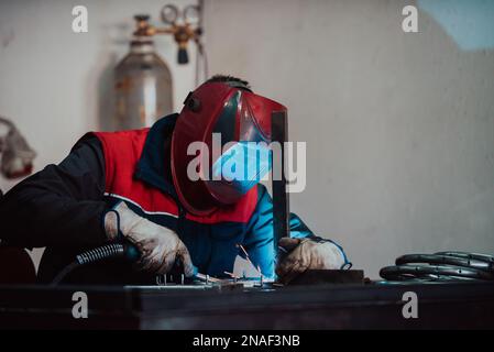 Saldatrice professionale in uniforme protettiva e maschera saldante tubo metallico sul tavolo industriale con altri lavoratori dietro nell'officina industriale. Foto Stock