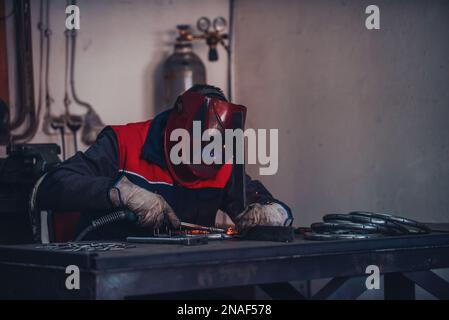 Saldatrice professionale in uniforme protettiva e maschera saldante tubo metallico sul tavolo industriale con altri lavoratori dietro nell'officina industriale. Foto Stock