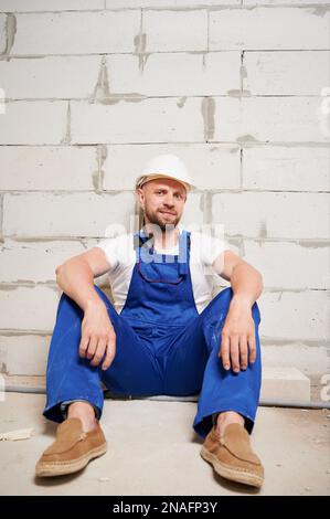 Costruttore maschile in tute da lavoro seduti sul pavimento in un edificio in costruzione o in ristrutturazione. Uomo bearded nel casco di sicurezza appoggiato contro la parete del mattone e guardando la macchina fotografica. Foto Stock