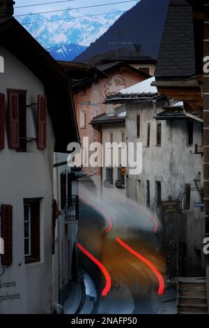 Le luci di posizione posteriori si sfocano mentre un autobus attraversa l'edificio lungo le strette strade del villaggio svizzero di Santa Maria. Il fascino delle comunità montane... Foto Stock
