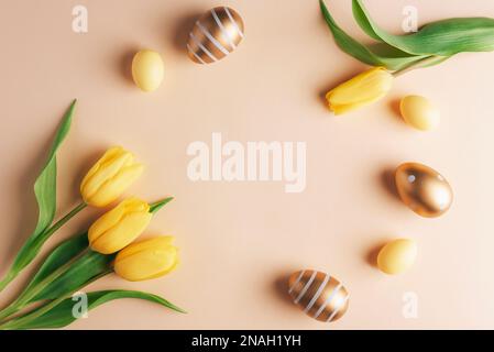 Cornice di uova pasquali e tulipani decorati in oro e giallo su sfondo beige neutro. Vista dall'alto, disposizione piatta, spazio di copia. Foto Stock