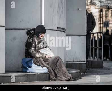 Bucarest, Romania - 12.29.2022: Ritratto di una donna anziana solitaria seduta sul marciapiede nel centro di Bucarest. Foto Stock