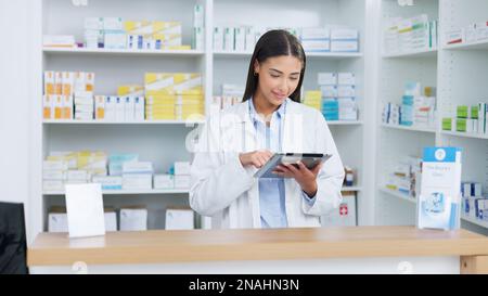 Ritratto di un farmacista allegro e amichevole utilizzando un tablet digitale per controllare l'inventario o gli ordini online in un chimico. Giovane donna latino usando Foto Stock