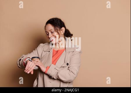 Bella donna che controlla il tempo sul suo orologio da polso, felice di essere puntuale per una riunione, isolato su sfondo beige Foto Stock